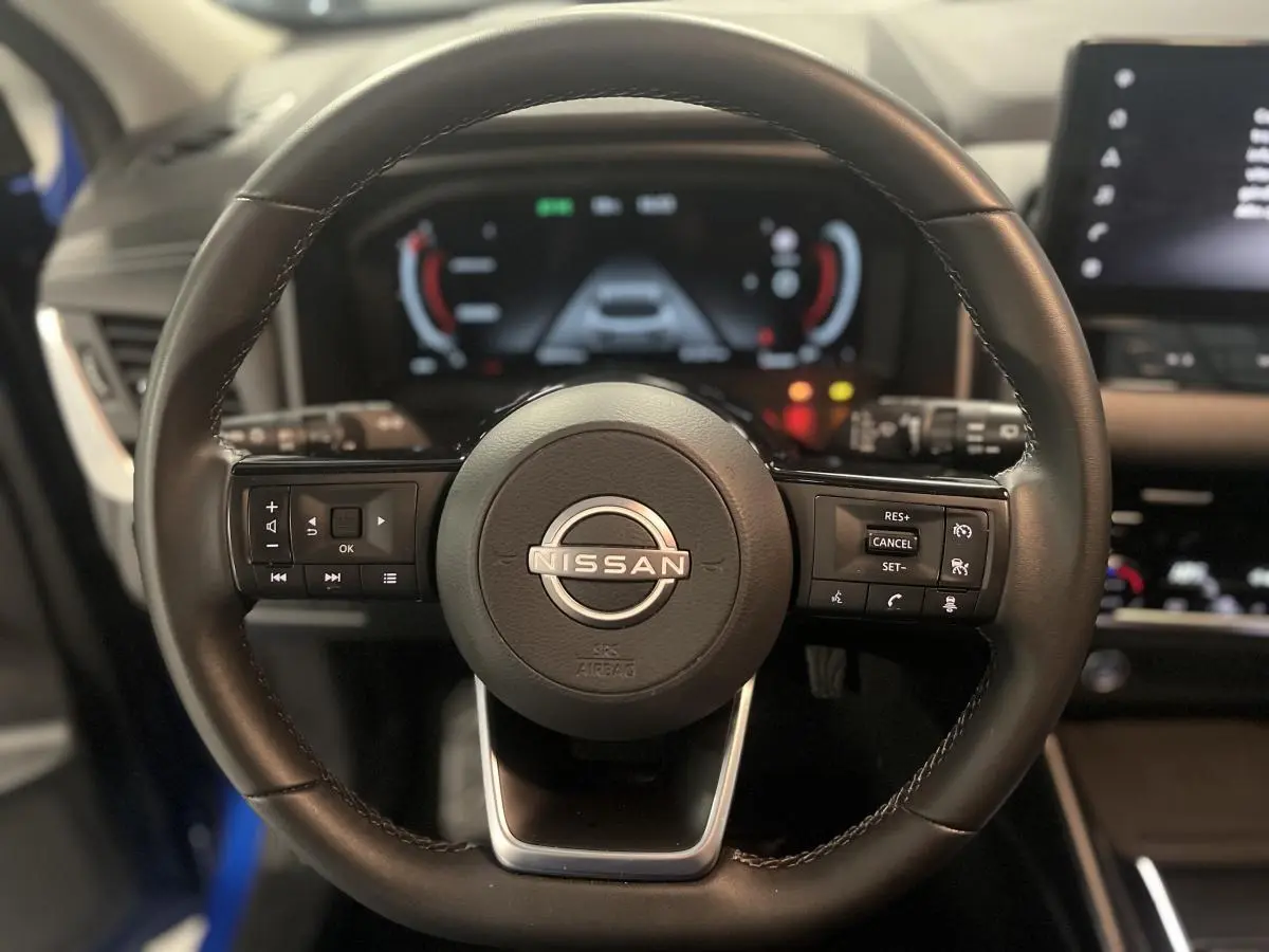 Volant cuir noir de Nissan Qashqai 1.3 Mild Hybrid 140 Shadow avec commandes intégrées et tableau de bord numérique flou.