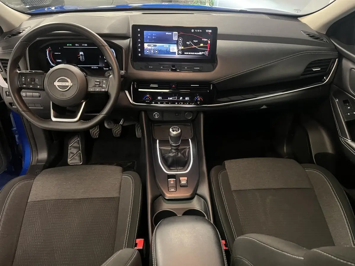 Intérieur avant du Nissan Qashqai 2024 bleu, vue du tableau de bord avec écran tactile et levier de vitesses manuel.