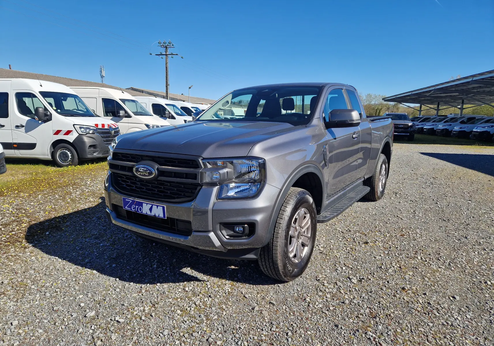 Ford Ranger Super Cabine gris carbone vu de 3/4 avant droit sur un parking en extérieur, avec calandre noire distinctive.