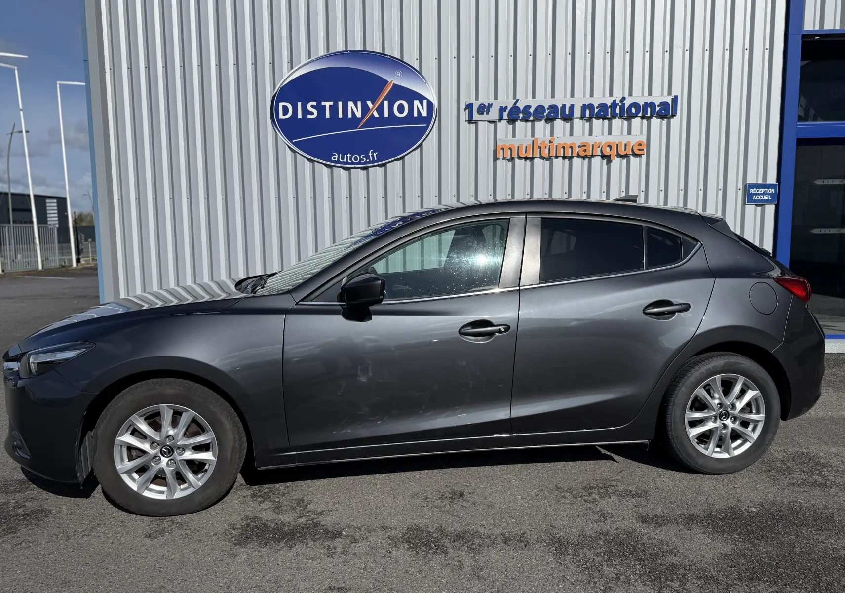 Vue de profil côté gauche d'une Mazda3 gris foncé stationnée devant un bâtiment avec un panneau Distinxion.