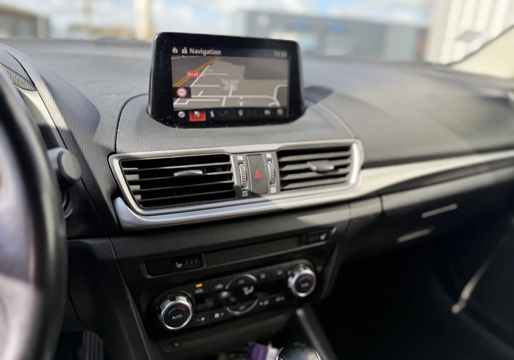 Vue intérieure centrée sur le tableau de bord de la Mazda3 2017, écran navigation et commandes climatisation visibles.