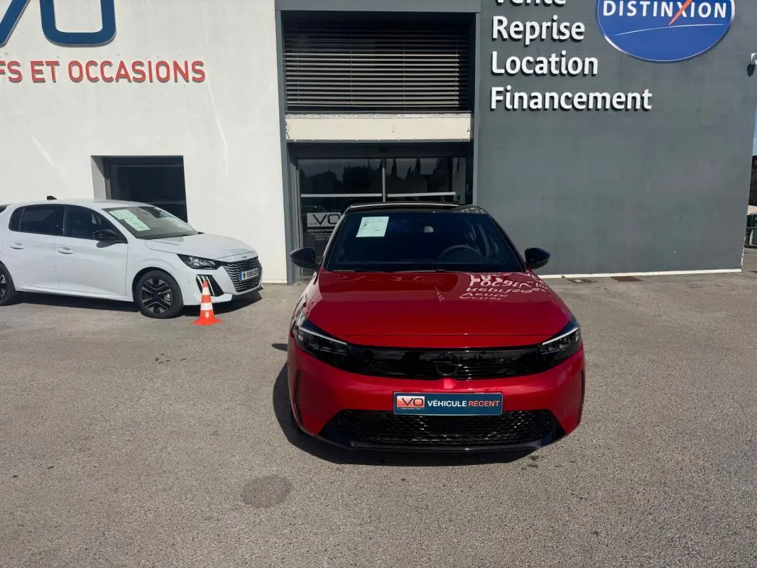 Vue avant d'une Opel Corsa rouge 2025 garée devant un bâtiment avec un panneau publicitaire visible.