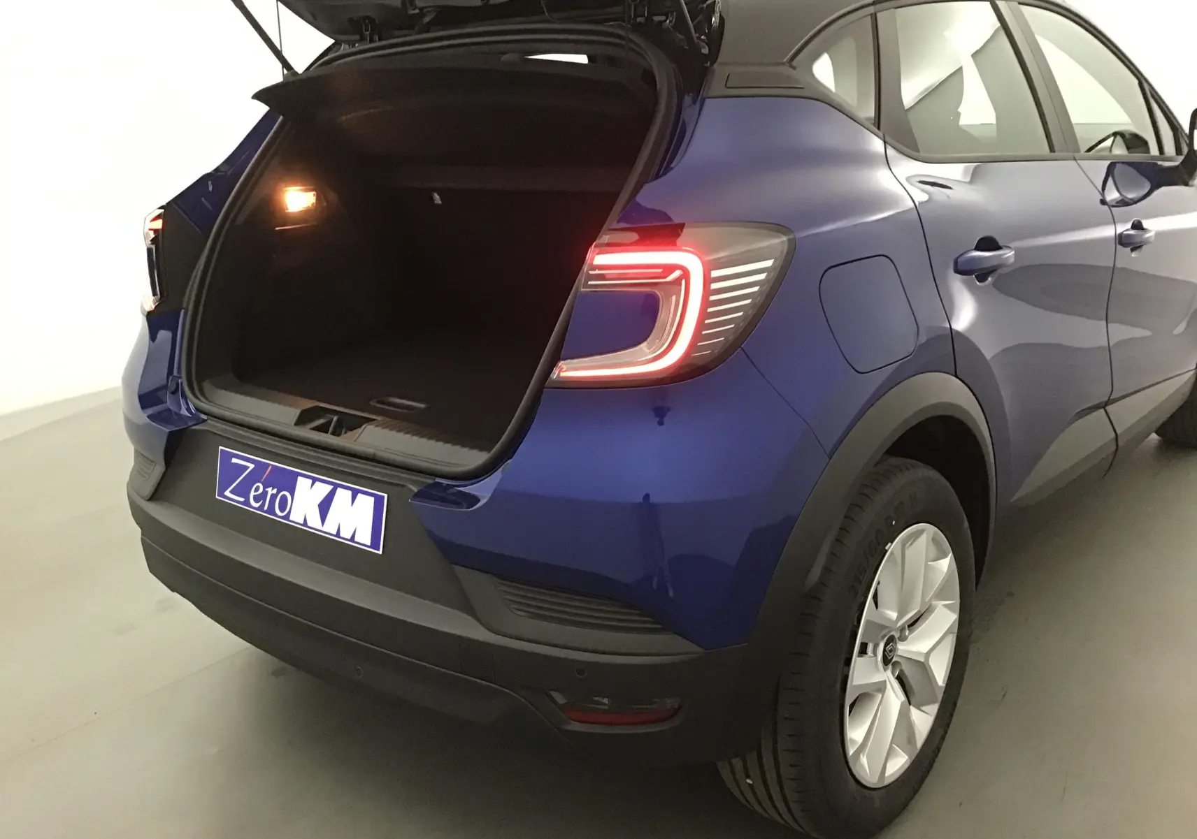 Vue 3/4 arrière droit du Renault Captur bleu iron/noir 2025 avec coffre ouvert et feux arrière allumés.