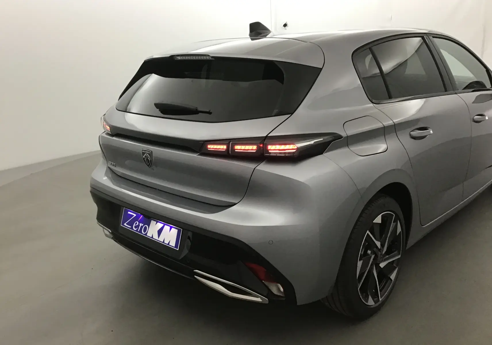 Vue 3/4 arrière droite d'une Peugeot 308 gris Artense avec feux arrière allumés et jantes alliage noires.