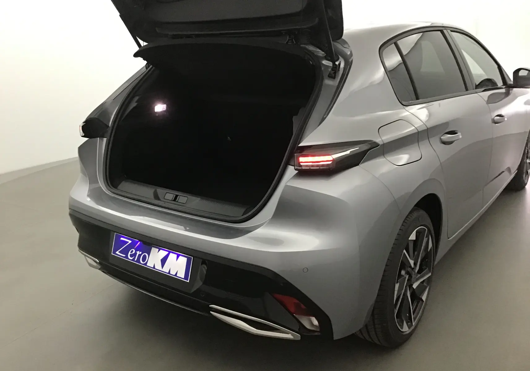 Vue 3/4 arrière droit d'une Peugeot 308 gris Artense avec coffre ouvert et feux arrière allumés dans un studio.