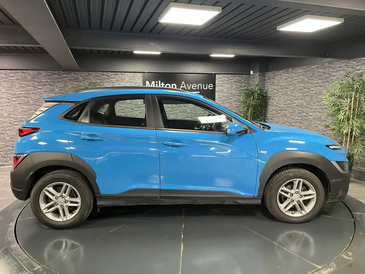Hyundai Kona bleu en profil côté gauche, avec protections noires et jantes alliage 16 pouces visibles en intérieur showroom.