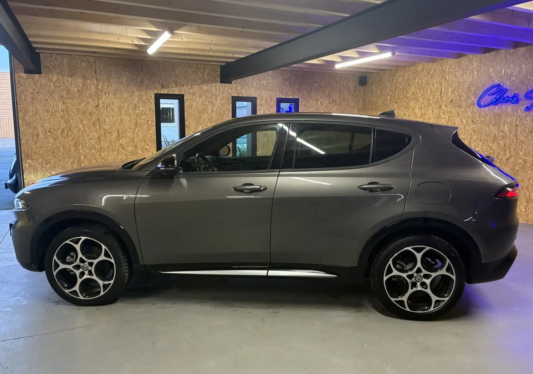 Vue de profil côté gauche d'un Alfa Romeo Tonale gris foncé hybride rechargeable, dans un garage avec mur en bois.