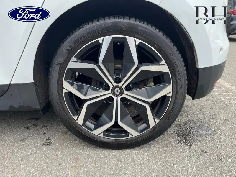 Gros plan sur la roue avant droite d’un Renault Scénic blanc nacré avec jante alliage noire et argentée au design géométrique.