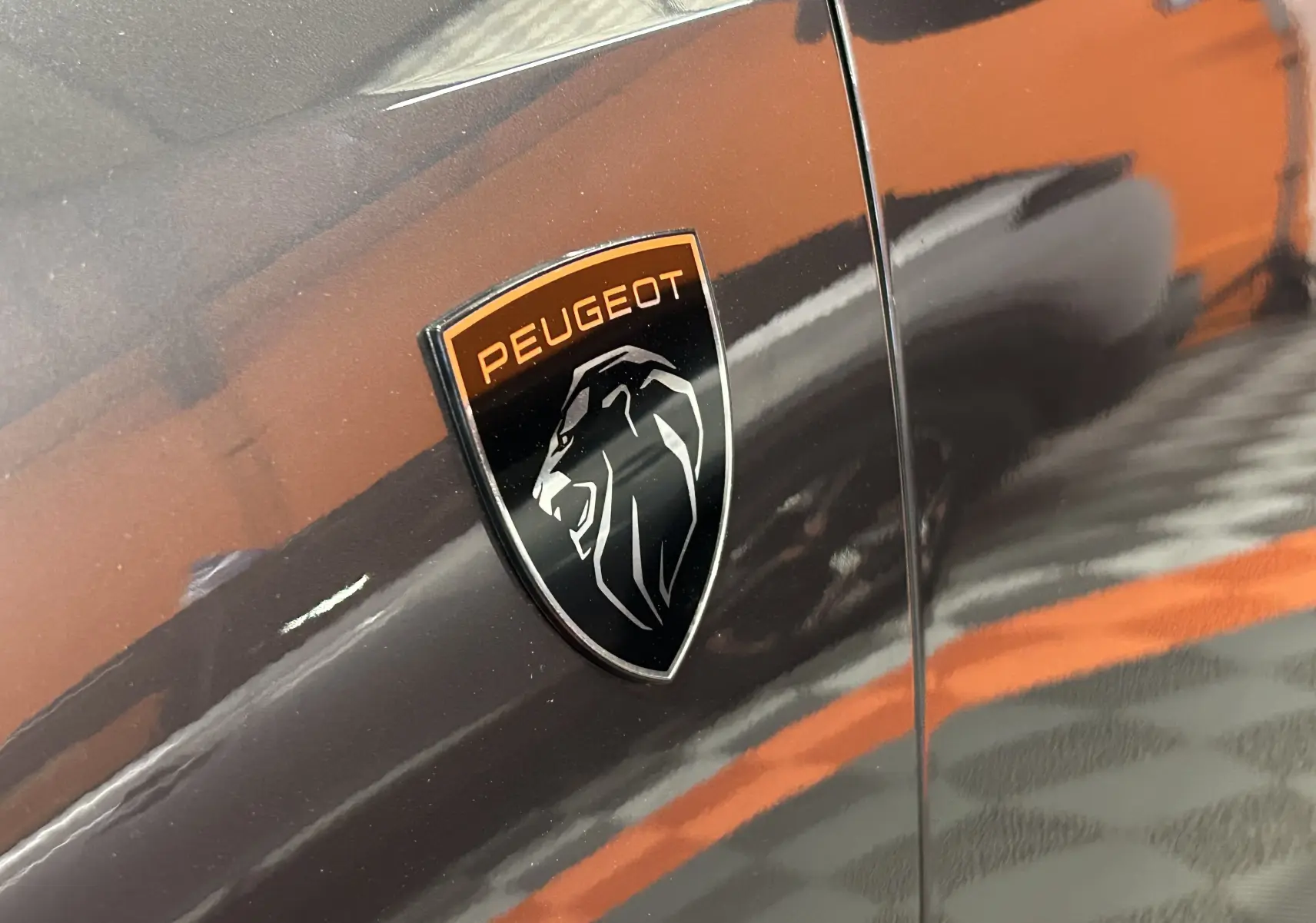 Gros plan sur l'écusson Peugeot noir et orange sur la porte gris titane du SUV 5008 Hybrid 2025.