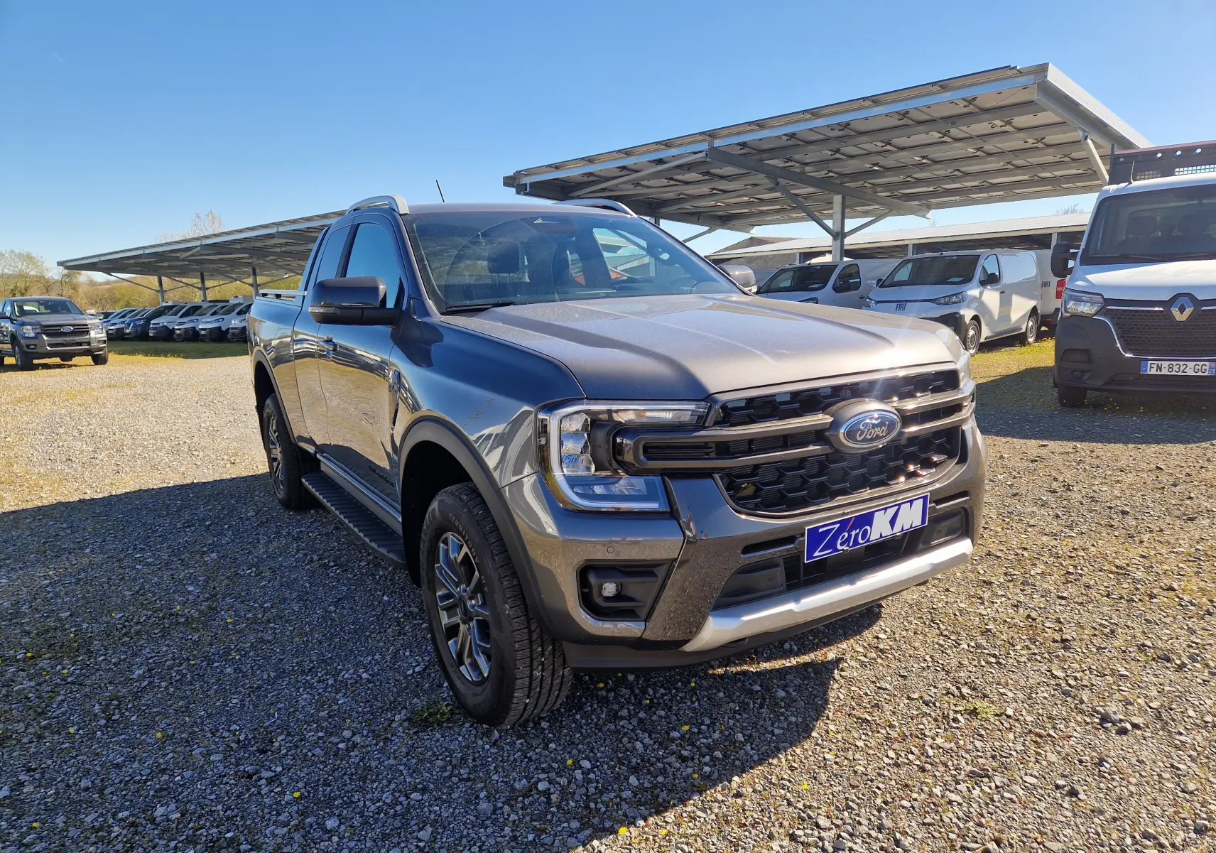 Vue 3/4 avant droit d'un Ford Ranger Super Cabine gris carbone avec calandre noire et plaques "Zero KM" sur un parking extérieur.