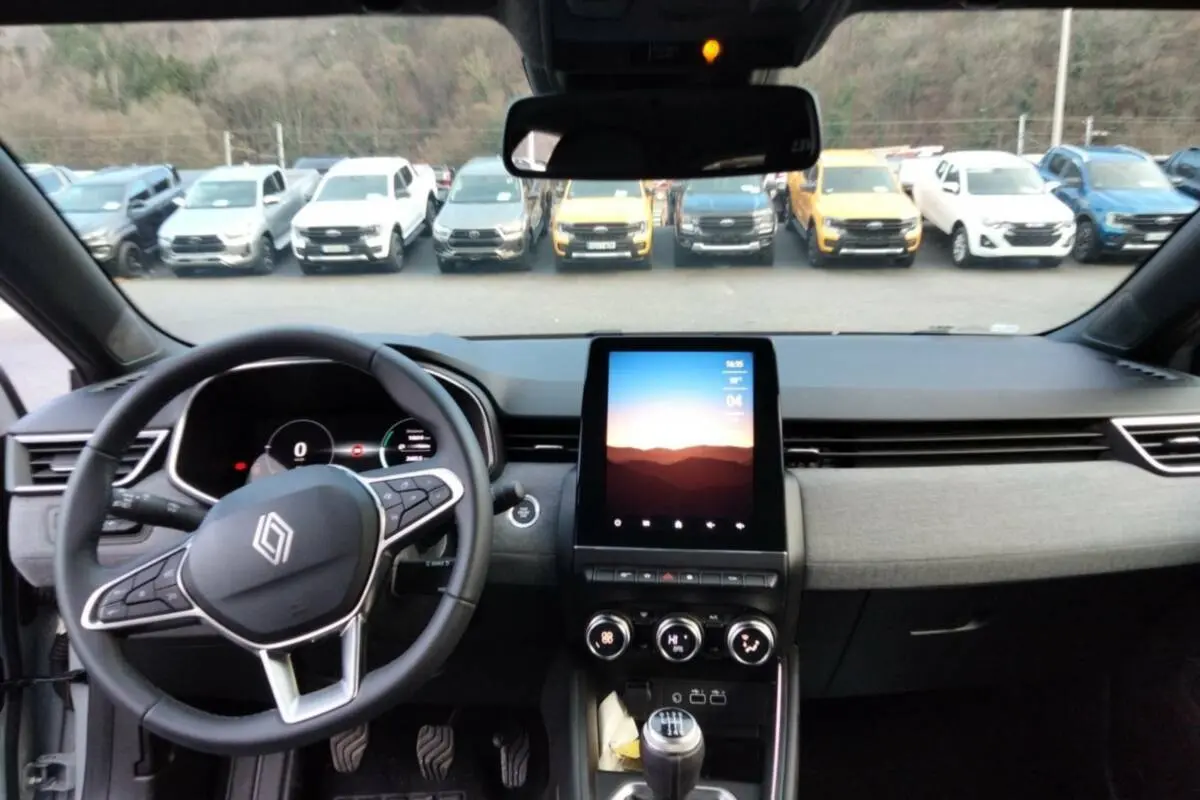 Vue intérieure avant de la Renault Clio V TCe 90 Techno Plus 2025, tableau de bord gris avec écran tactile vertical et volant multifonctions.