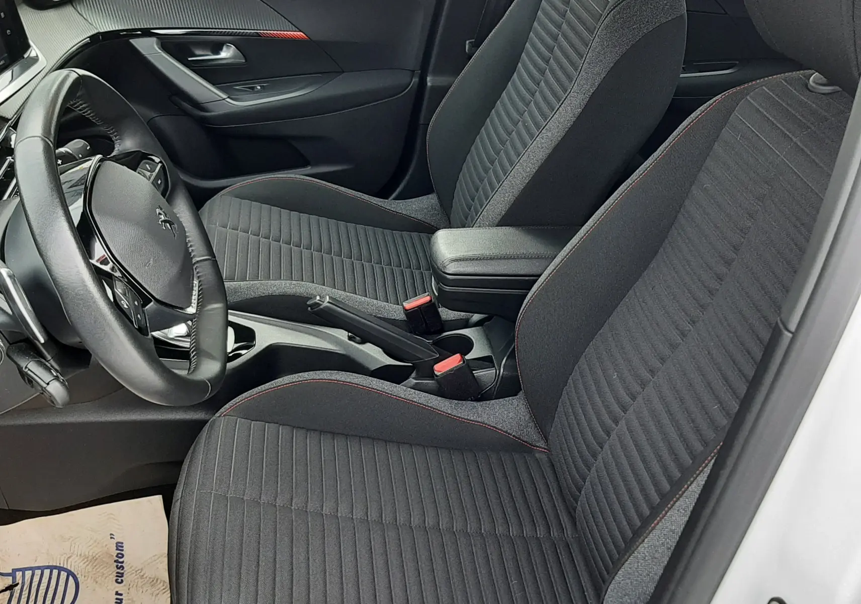 Vue intérieure côté conducteur du Peugeot 2008 blanc 2021, sièges tissu noir avec surpiqûres rouges et volant cuir.
