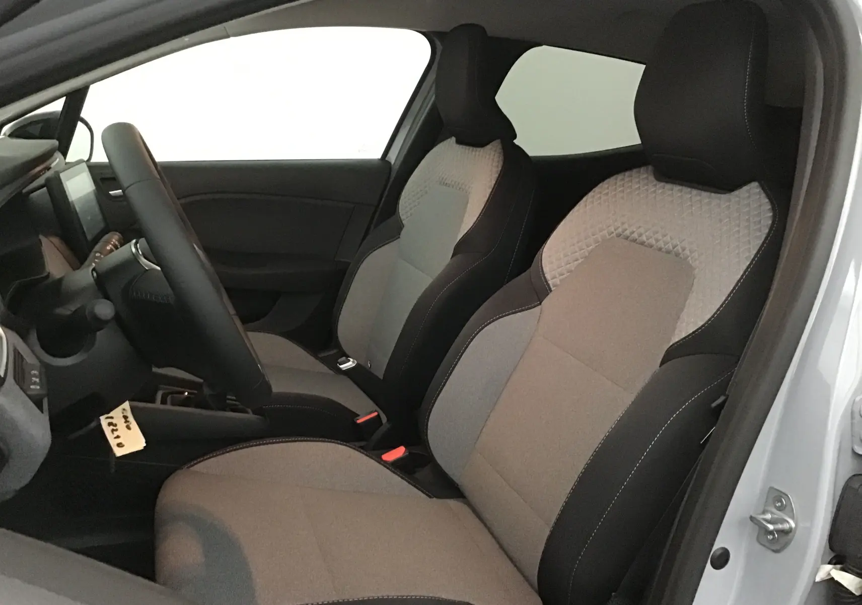 Vue intérieure côté conducteur de la Renault Clio V 2025, sièges tissu gris clair et noir avec surpiqûres contrastées.