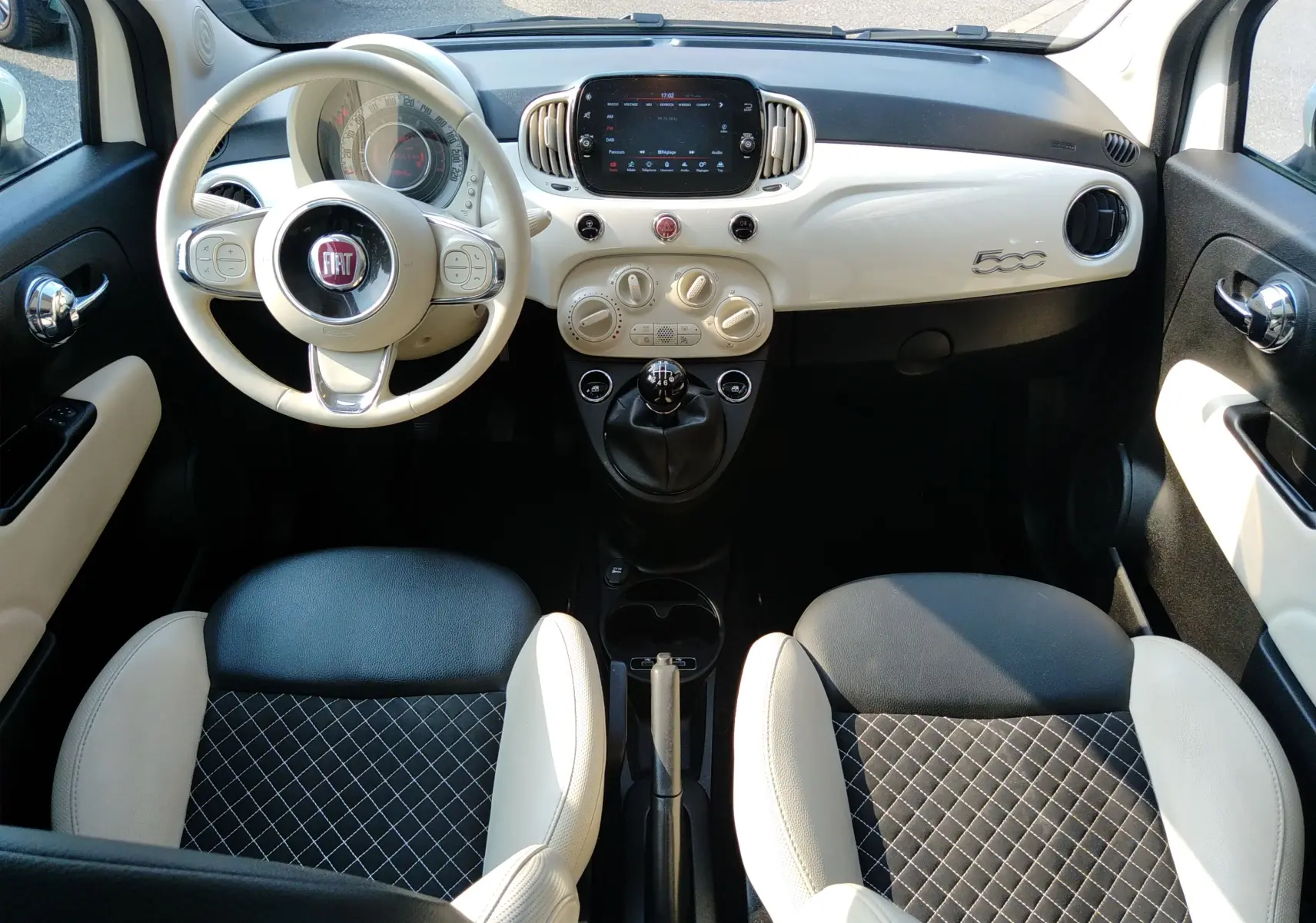 Vue intérieure avant de la Fiat 500 blanche, avec volant beige, tableau de bord épuré et sièges bi-color noir et beige.