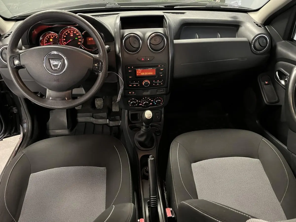 Vue intérieure avant du Dacia Duster noir 2016, volant, tableau de bord et sièges bicolores gris et noir visibles.