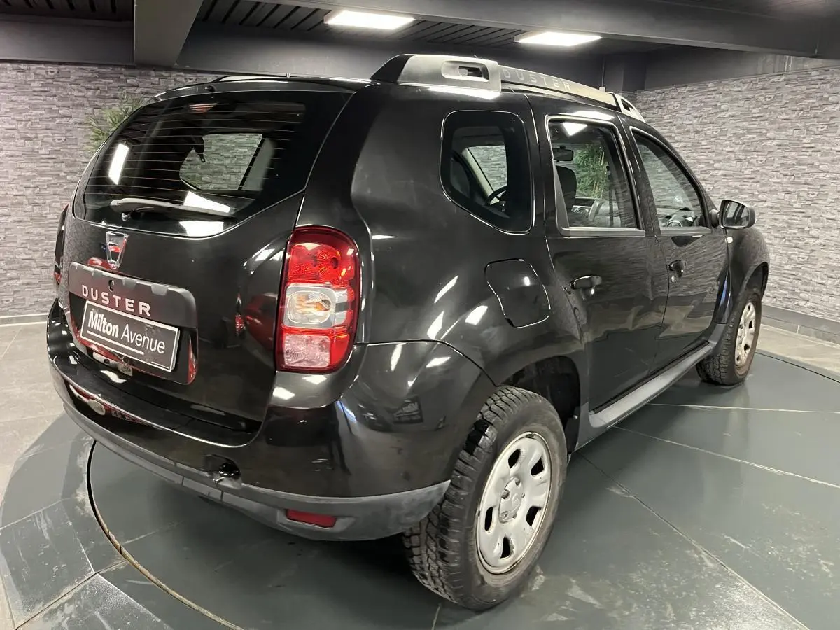 Vue 3/4 arrière droite d'un Dacia Duster noir avec barres de toit et jantes acier en intérieur showroom.