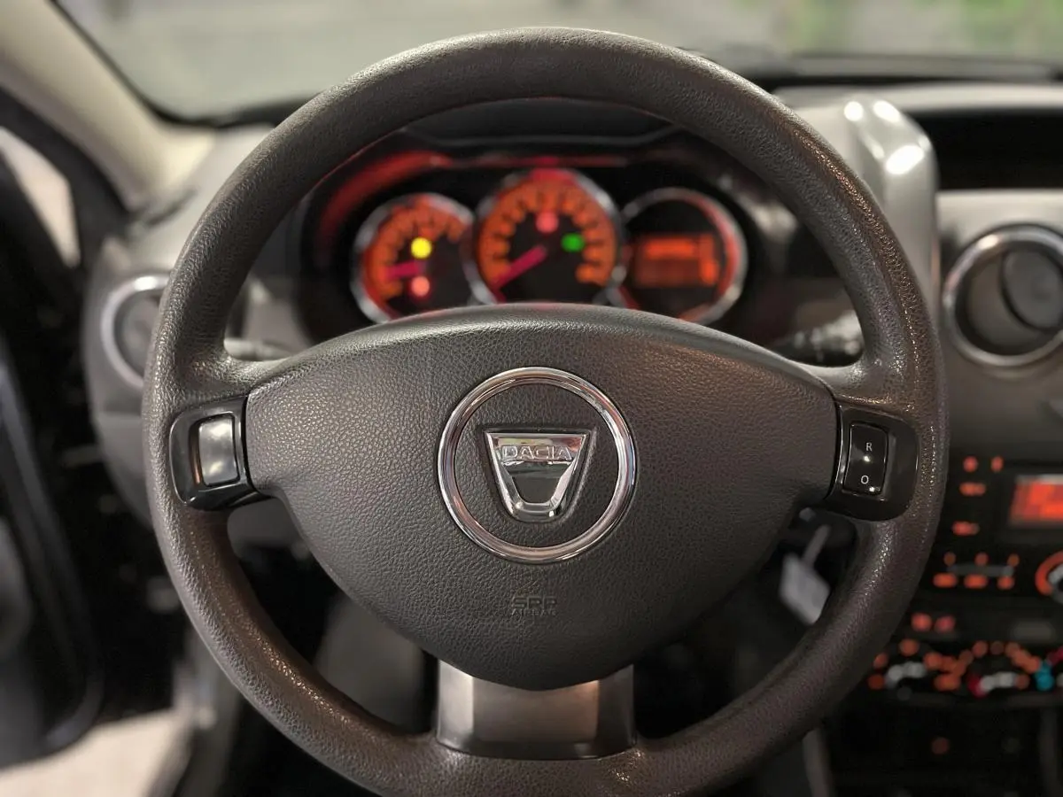 Volant noir avec logo chromé Dacia au centre, tableau de bord éclairé en orange flou en arrière-plan.