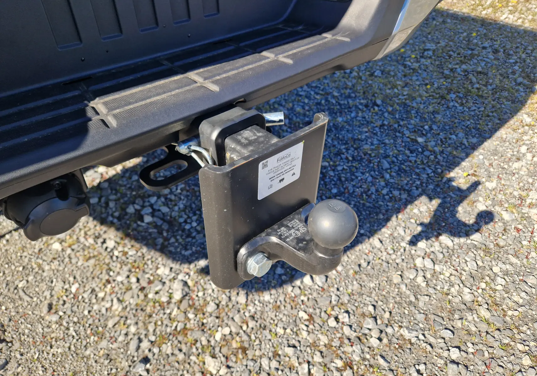 Gros plan sur l'attelage arrière noir du Ford Ranger Super Cabine gris Iconic, avec prise électrique visible sur sol gravillonné.