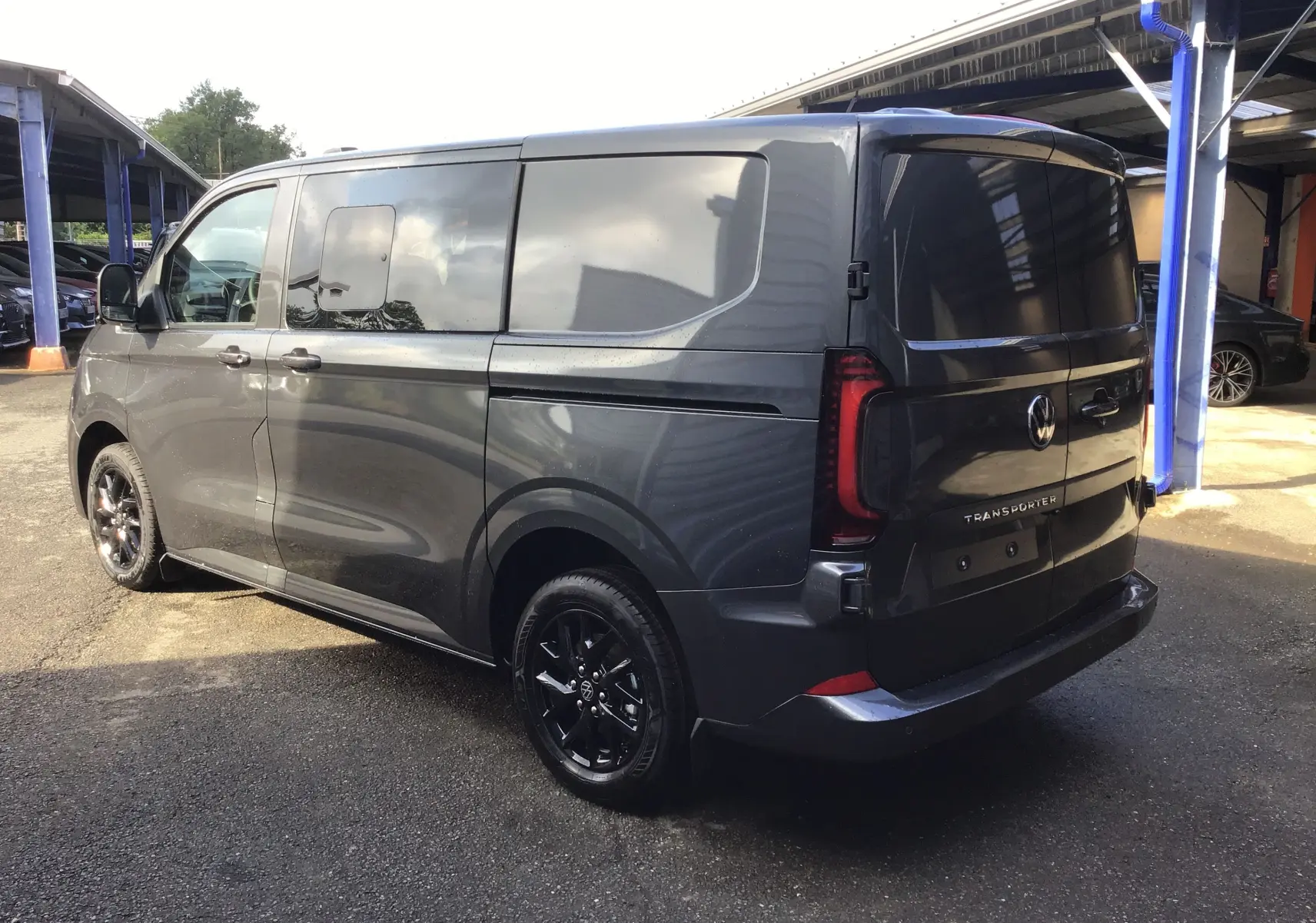 Vue 3/4 arrière droit d’un Volkswagen Transporter ProCab gris anthracite avec jantes alu noires diamantées sous abri.