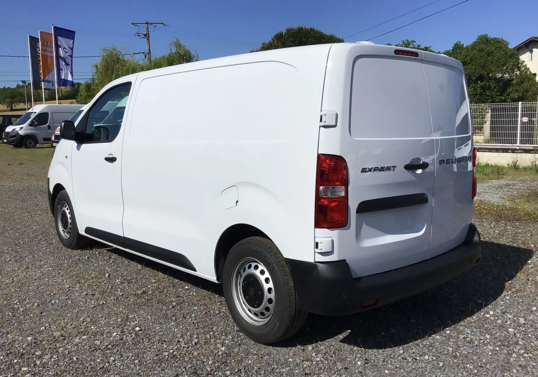 Vue 3/4 arrière droite d'un Peugeot Expert Fourgon blanc 2025 avec logo et feux arrière visibles sur un sol gravillonné.