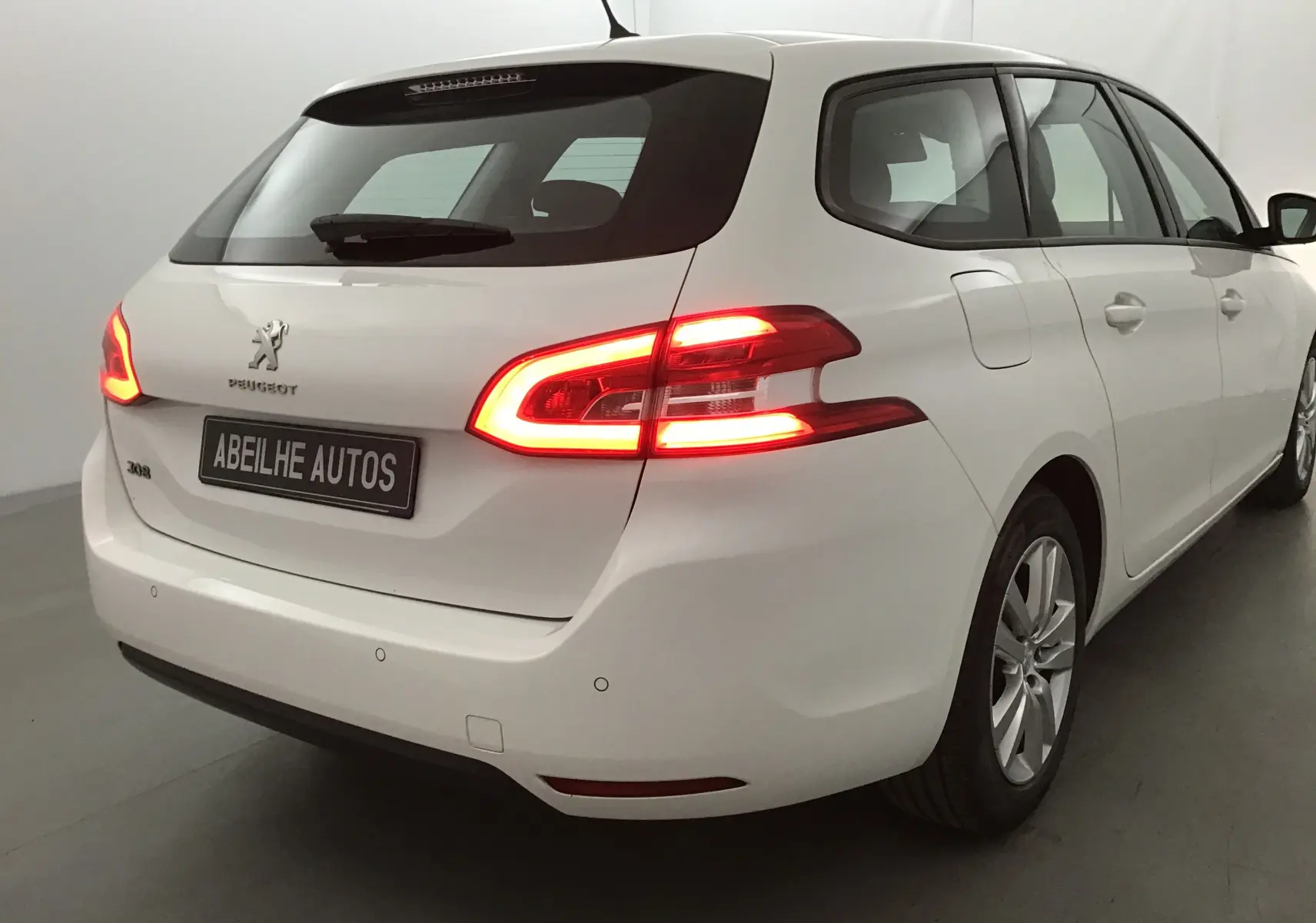 Vue 3/4 arrière droite d'une Peugeot 308 SW blanche de 2018 avec feux arrière allumés dans un studio.