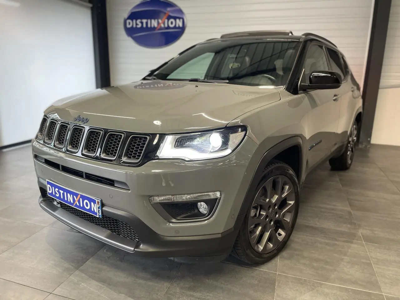 Jeep Compass gris foncé vu en 3/4 avant droit avec phares allumés et jantes alliage sombres.