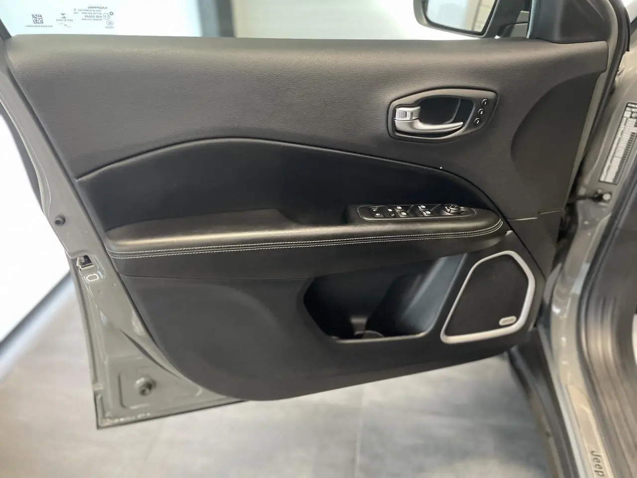 Porte avant gauche ouverte du Jeep Compass gris foncé, montrant garniture noire avec surpiqûres blanches et haut-parleur intégré.
