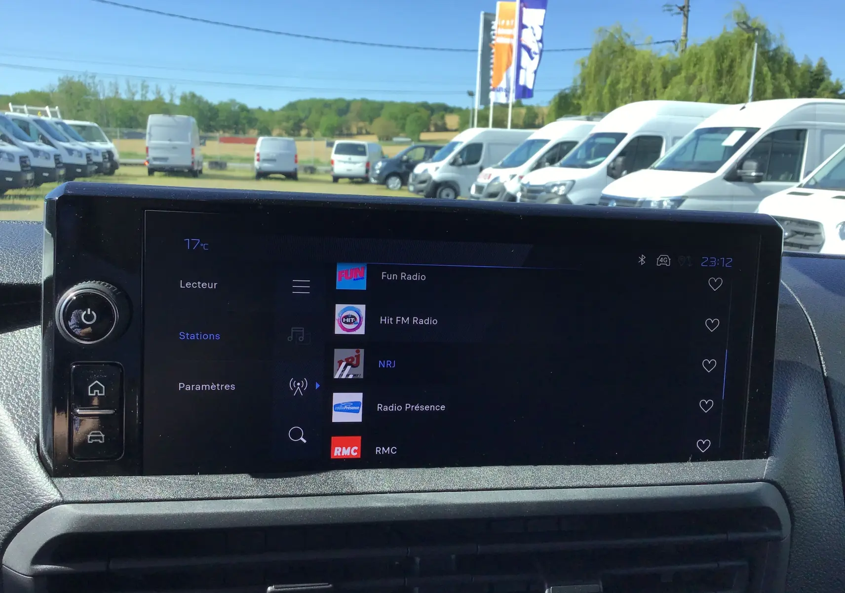 Écran tactile central du Peugeot Expert Fourgon blanc 2025 affichant les stations radio avec vue sur plusieurs fourgons blancs en arrière-plan.