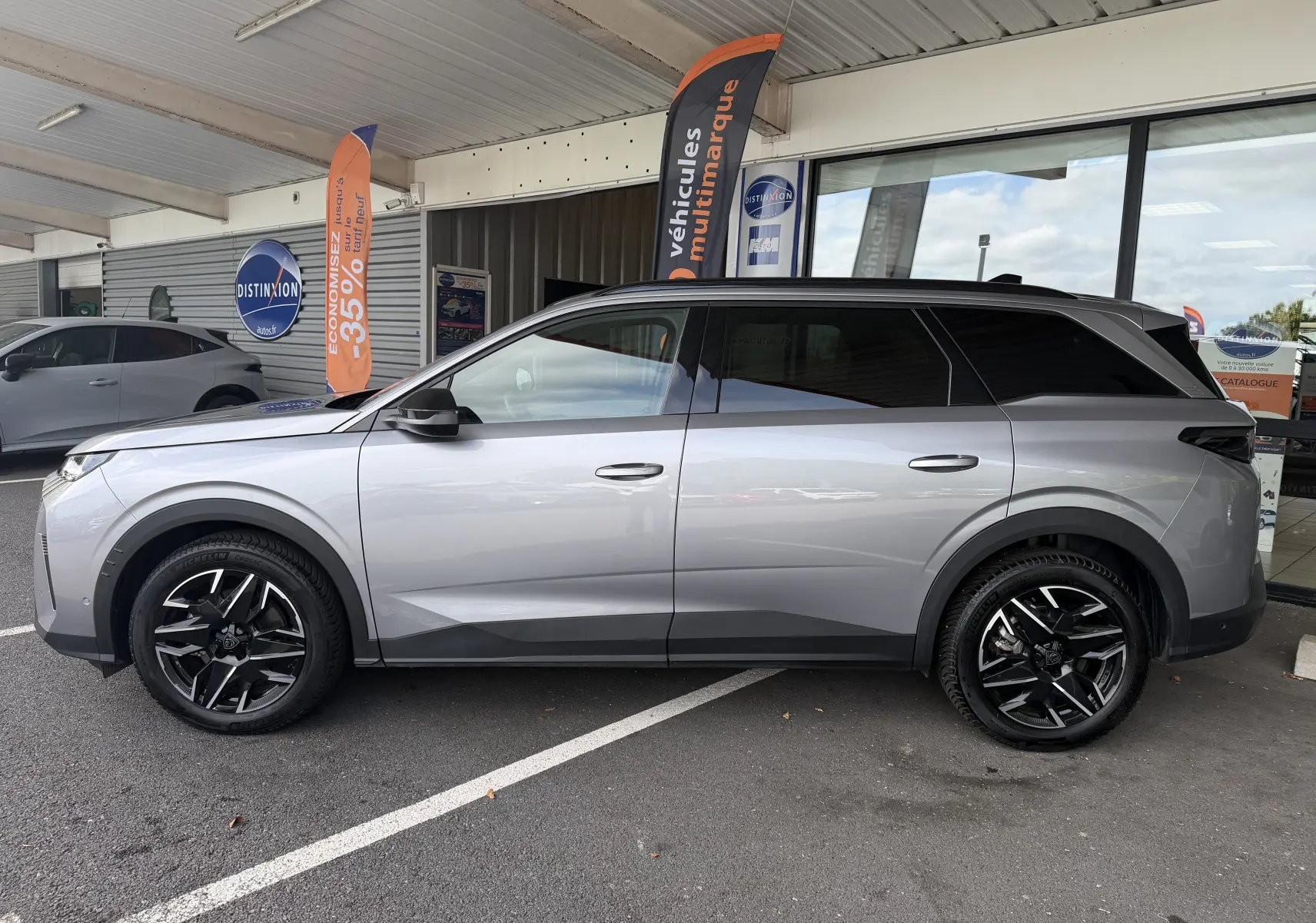 Vue de profil côté gauche d’un Peugeot 1007 gris artense avec jantes aluminium noires et toit noir, stationné devant un showroom.