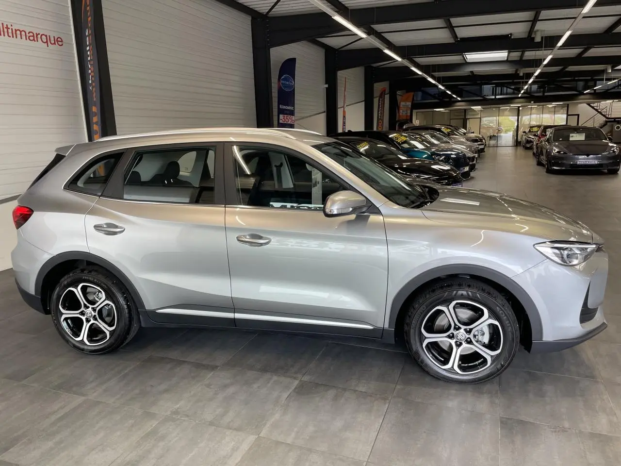 Vue de profil côté gauche du SUV MG ZS 1.5 Hybrid+ gris clair avec jantes alliage 17 pouces en showroom.