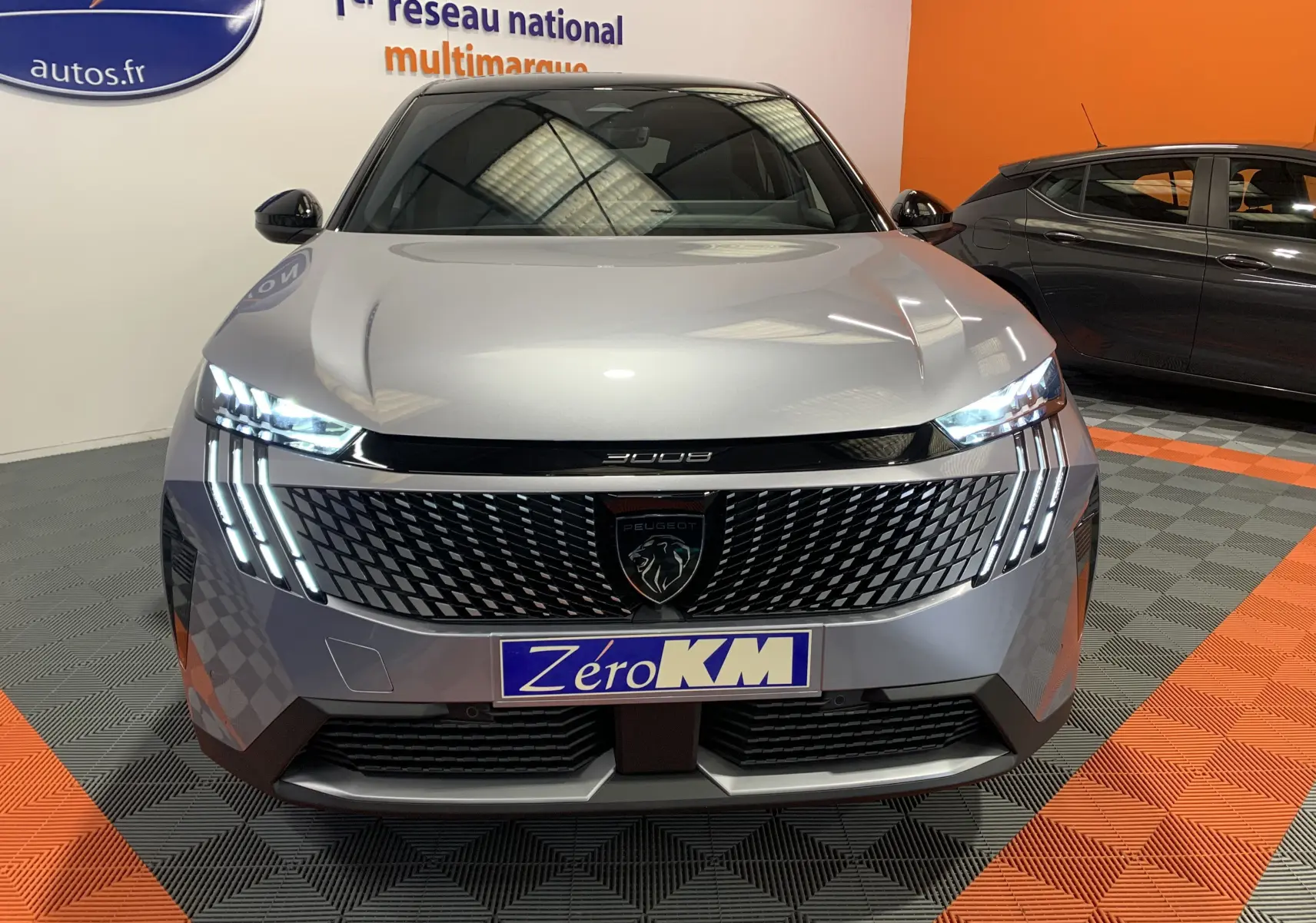 Vue frontale du Peugeot 3008 III GT Hybrid 136 e-DSC6 en gris Artense avec phares LED allumés et calandre distinctive.