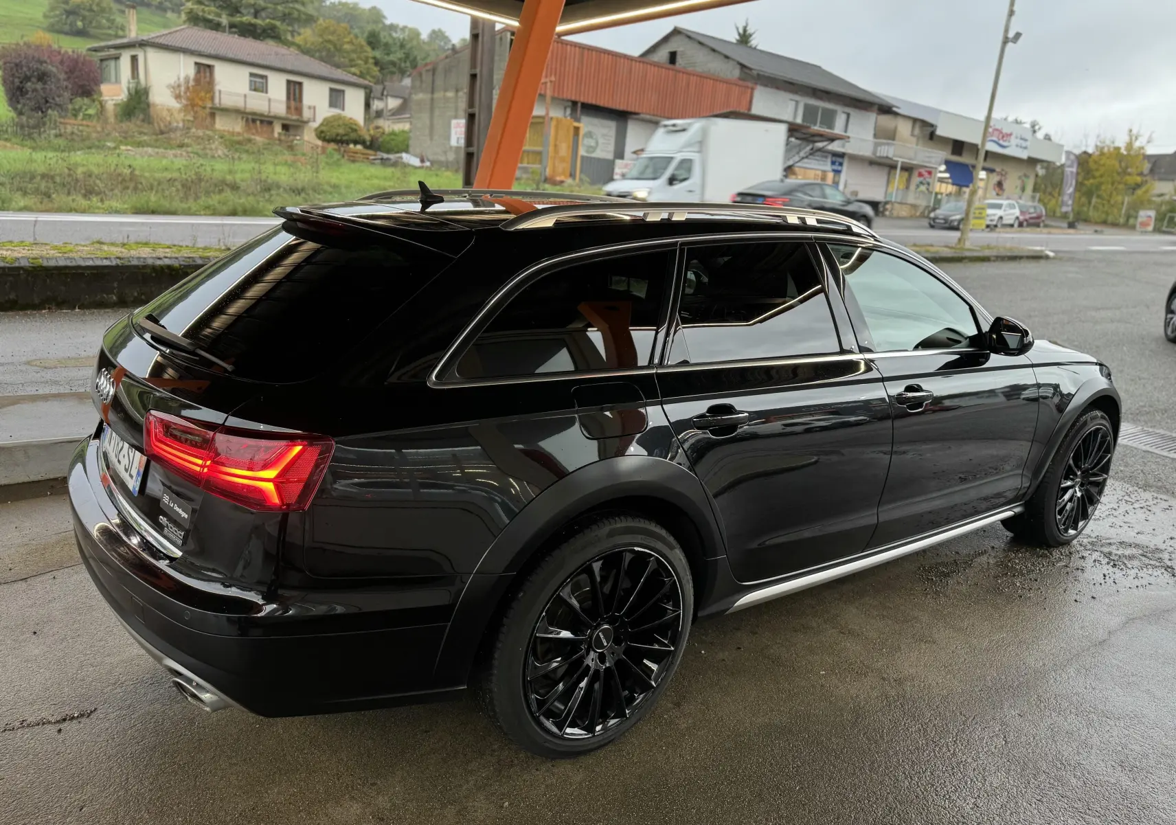 Audi A6 Avant Allroad noire vue 3/4 arrière droit, avec jantes noires et feux arrière allumés sous abri.