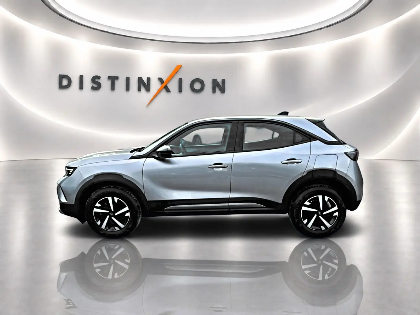 Opel Mokka gris en profil droit dans un showroom lumineux avec jantes noires et logo Distinxion en arrière-plan.