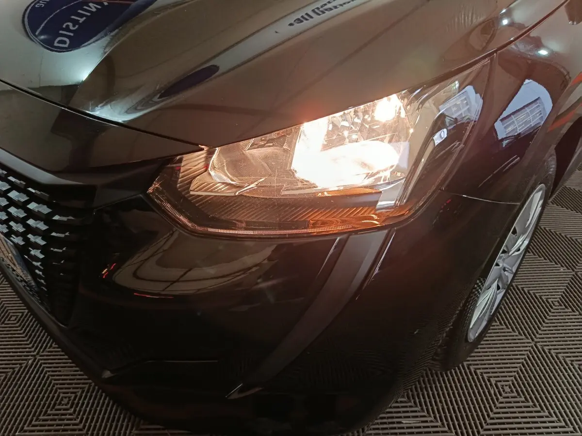 Gros plan sur le phare avant droit allumé d'une Peugeot 208 noire, mettant en valeur la calandre et la jante avant.