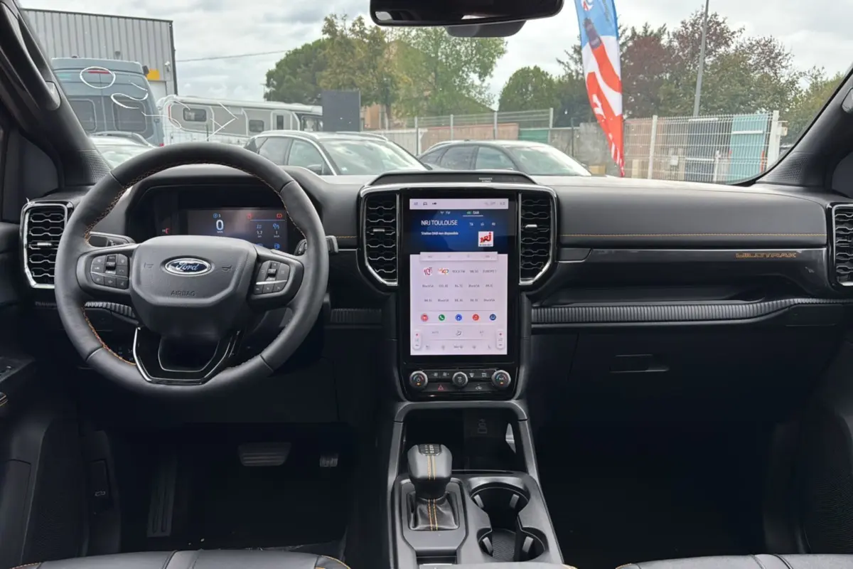 Vue intérieure frontale du Ford Ranger Wildtrak Plus 2025, tableau de bord noir avec écran tactile vertical et volant cuir multifonctions.