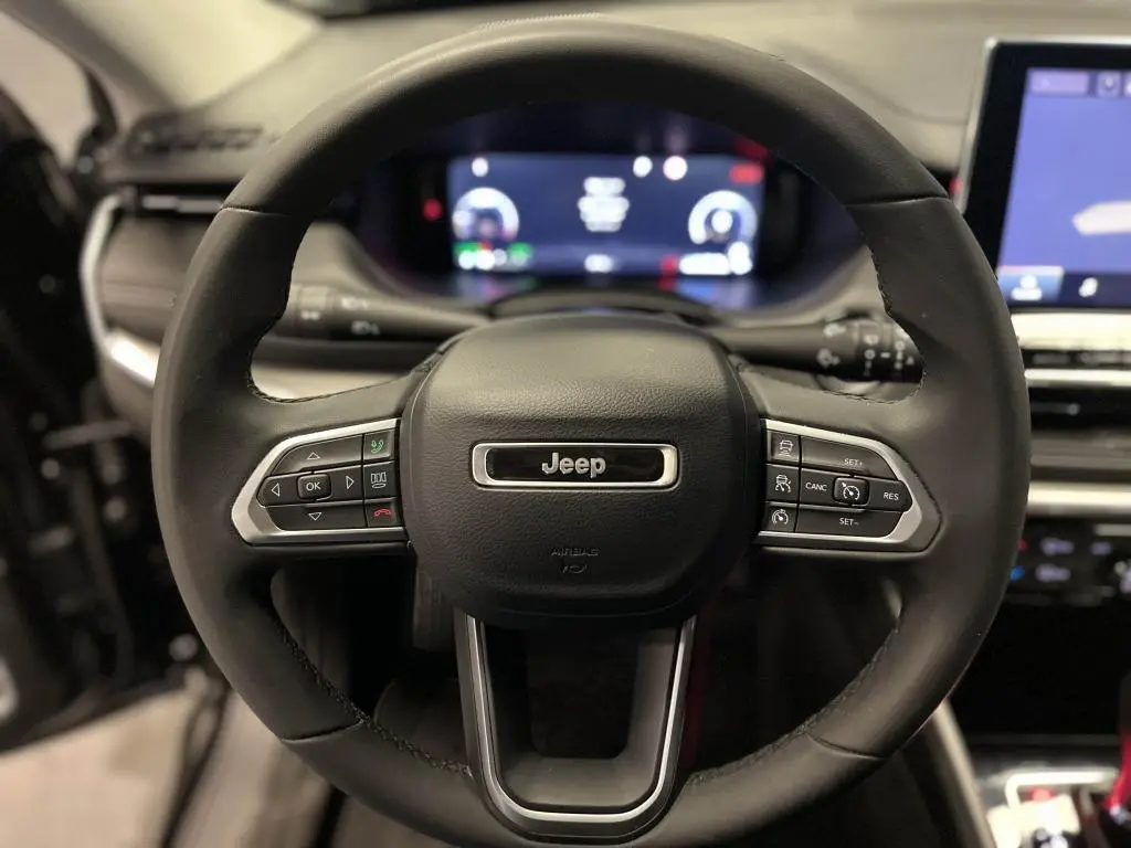 Volant en cuir noir du Jeep Compass 2024 avec commandes intégrées, tableau de bord numérique et écran tactile visible en arrière-plan.