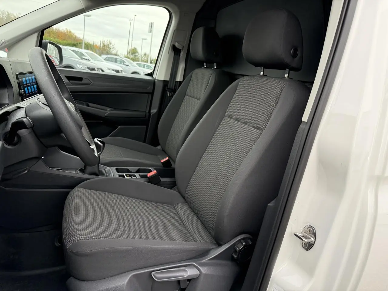 Vue intérieure côté conducteur du Volkswagen Caddy Maxi Cargo blanc candy, montrant les sièges avant en tissu gris et le volant multifonctions.