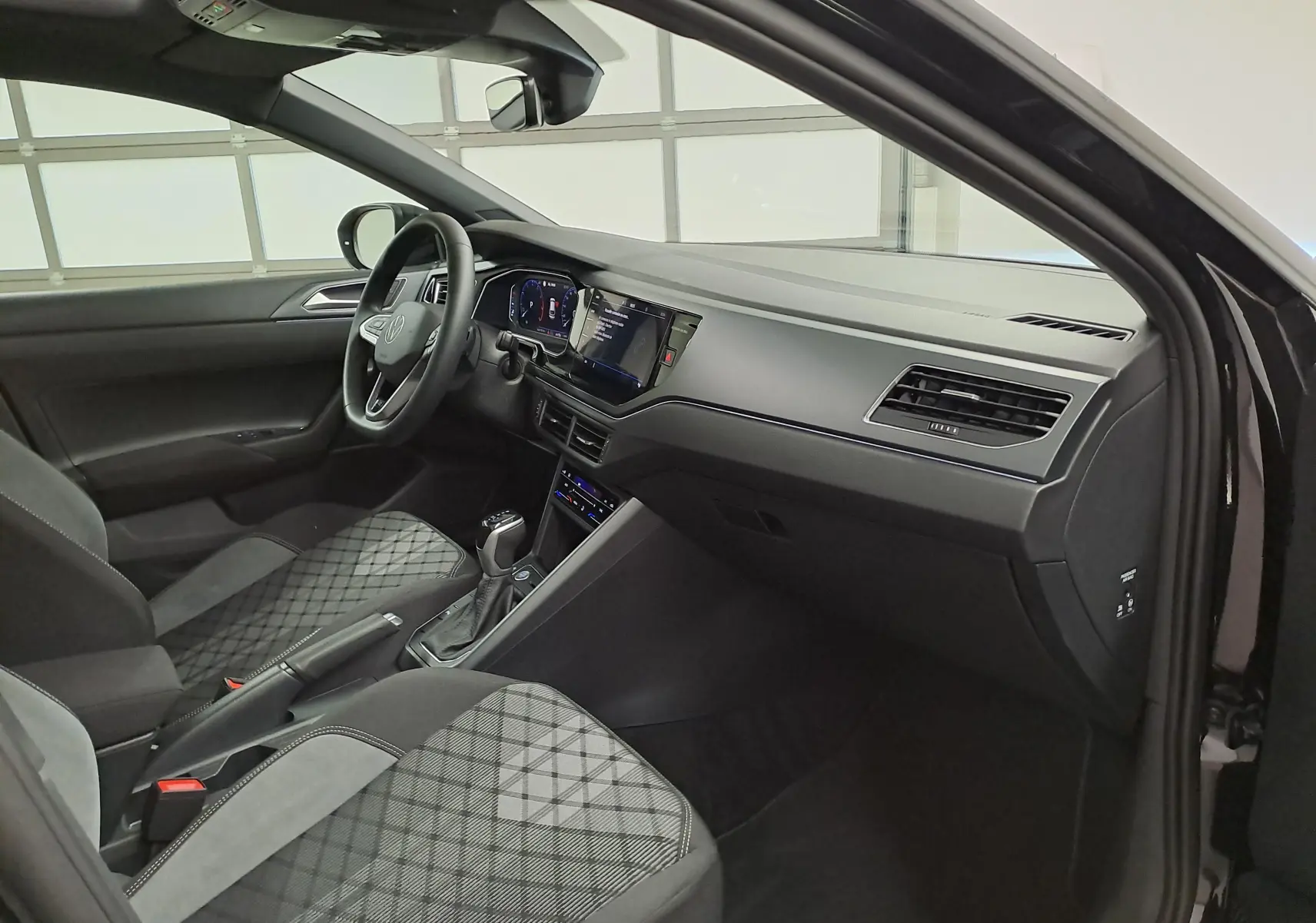 Vue intérieure côté passager avant du Volkswagen Taigo noir intense nacré, avec tableau de bord moderne et sièges tissu à motifs.