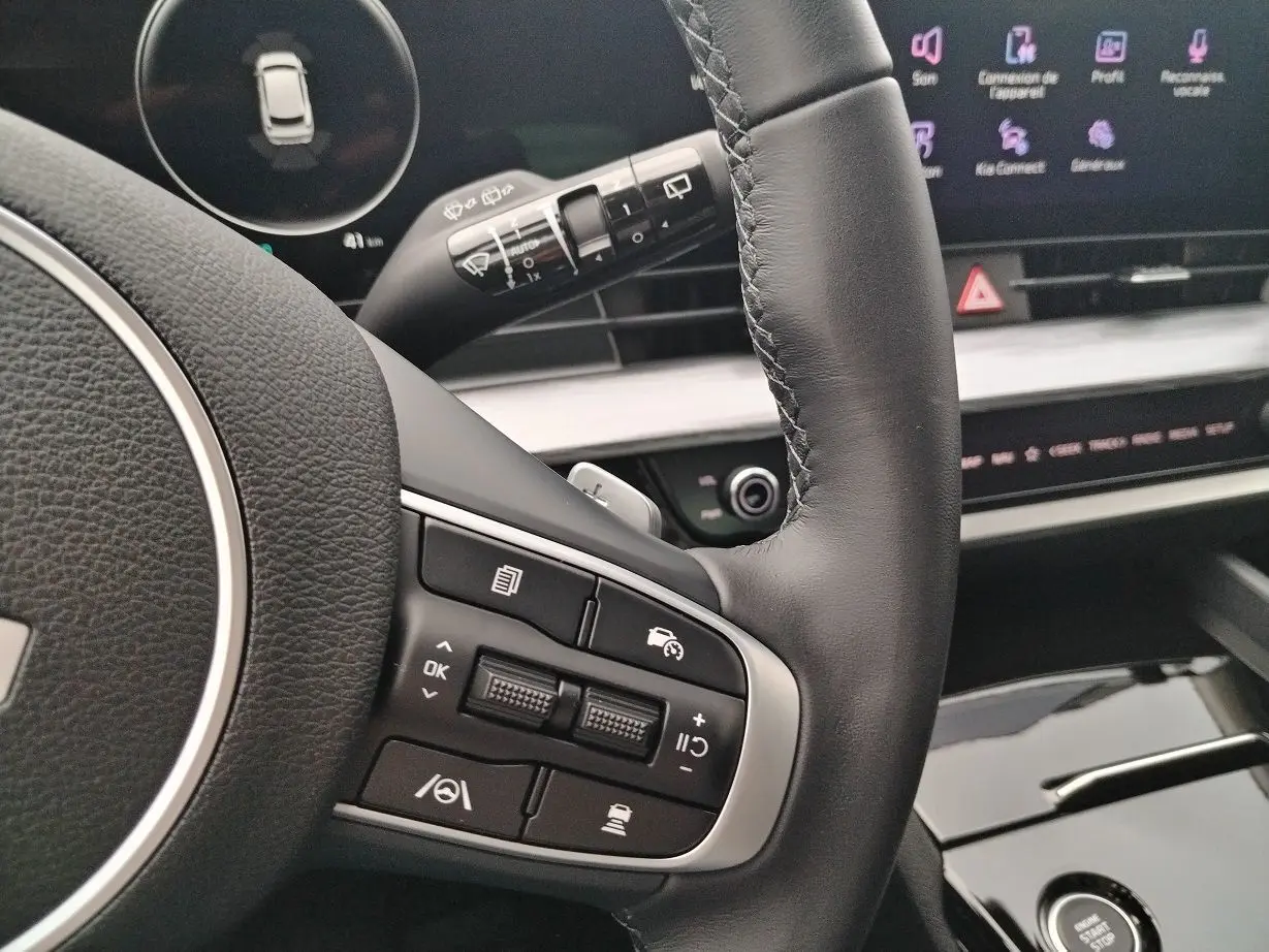 Détail du volant en cuir noir du KIA Sportage 2025, avec commandes intégrées et tableau de bord numérique en arrière-plan.