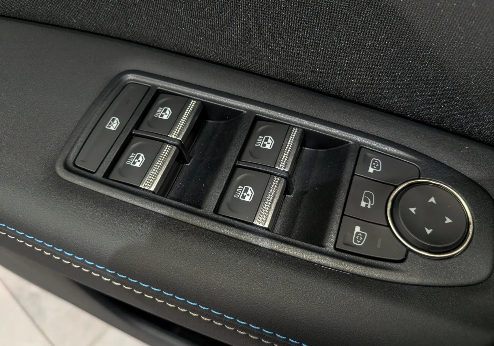 Gros plan sur les commandes de lève-vitres et réglages des rétroviseurs sur la porte côté conducteur, finition cuir noir avec surpiqûres bleues.