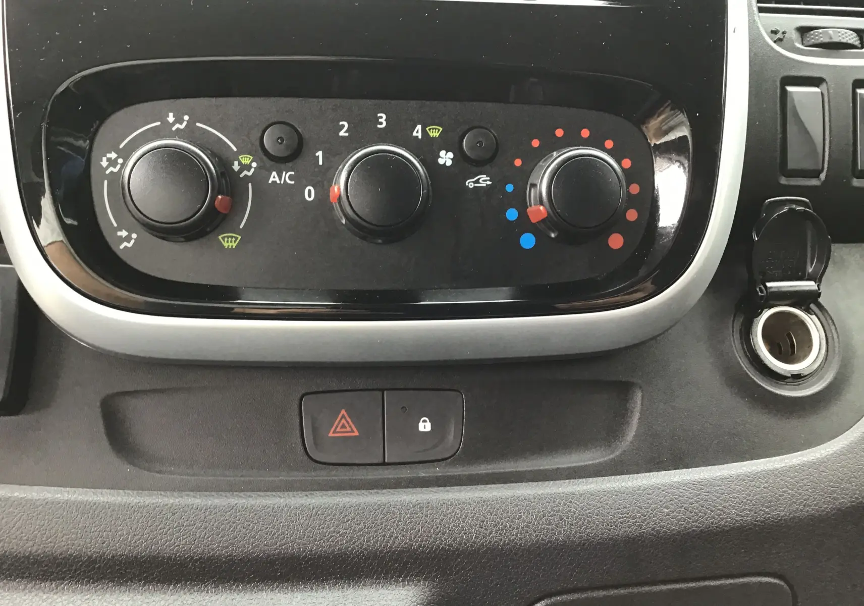 Gros plan sur les commandes de climatisation noires du Renault Trafic blanc, avec bouton d'alerte et prise allume-cigare visible.
