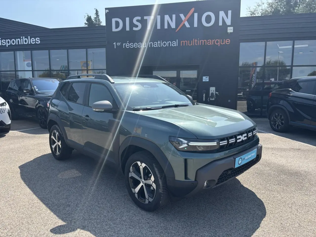 SUV Dacia Duster vert cèdre métal en 3/4 avant droit, avec jantes alliage et barres de toit visibles.