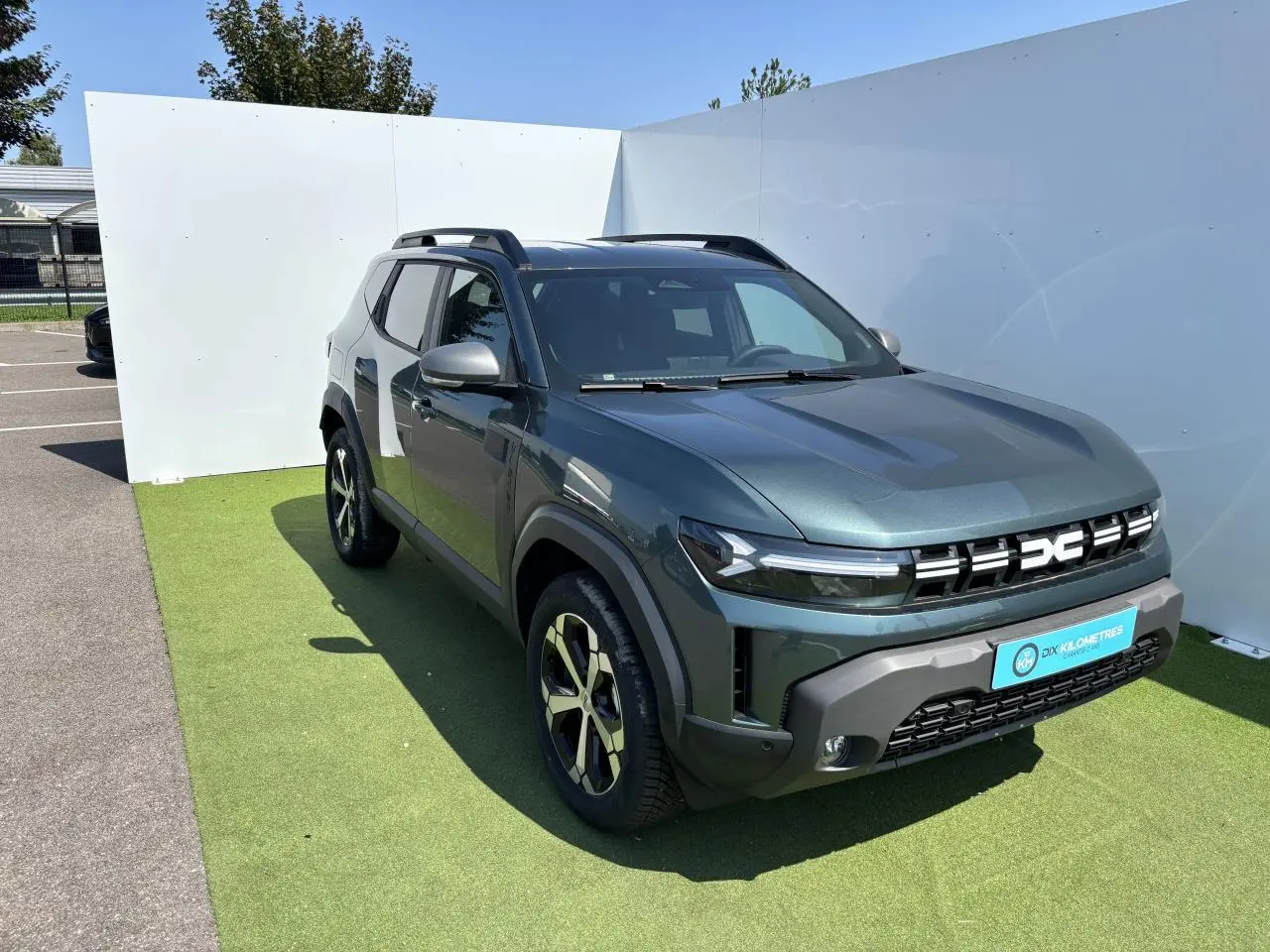 Dacia Duster III vert cèdre métal en 3/4 avant droit avec jantes alliage et barres de toit noires.