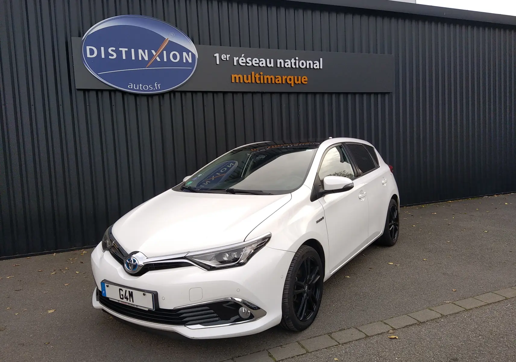 Toyota Auris hybride blanche vue en 3/4 avant droit, avec jantes noires et calandre chromée devant un mur noir.