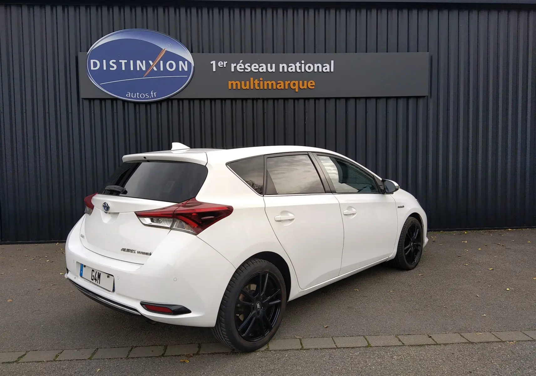 Toyota Auris hybride blanche vue 3/4 arrière droit, avec jantes noires et vitres teintées, stationnée devant un mur noir.