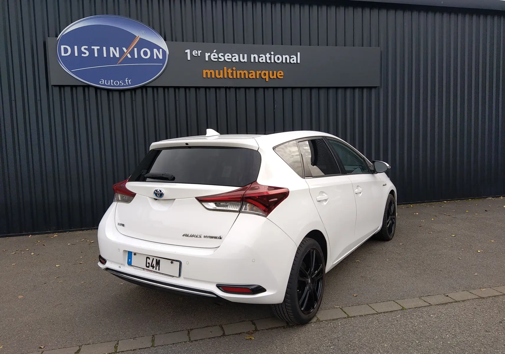 Toyota Auris hybride blanche vue 3/4 arrière droit, avec jantes noires et vitres teintées, stationnée devant un mur sombre.