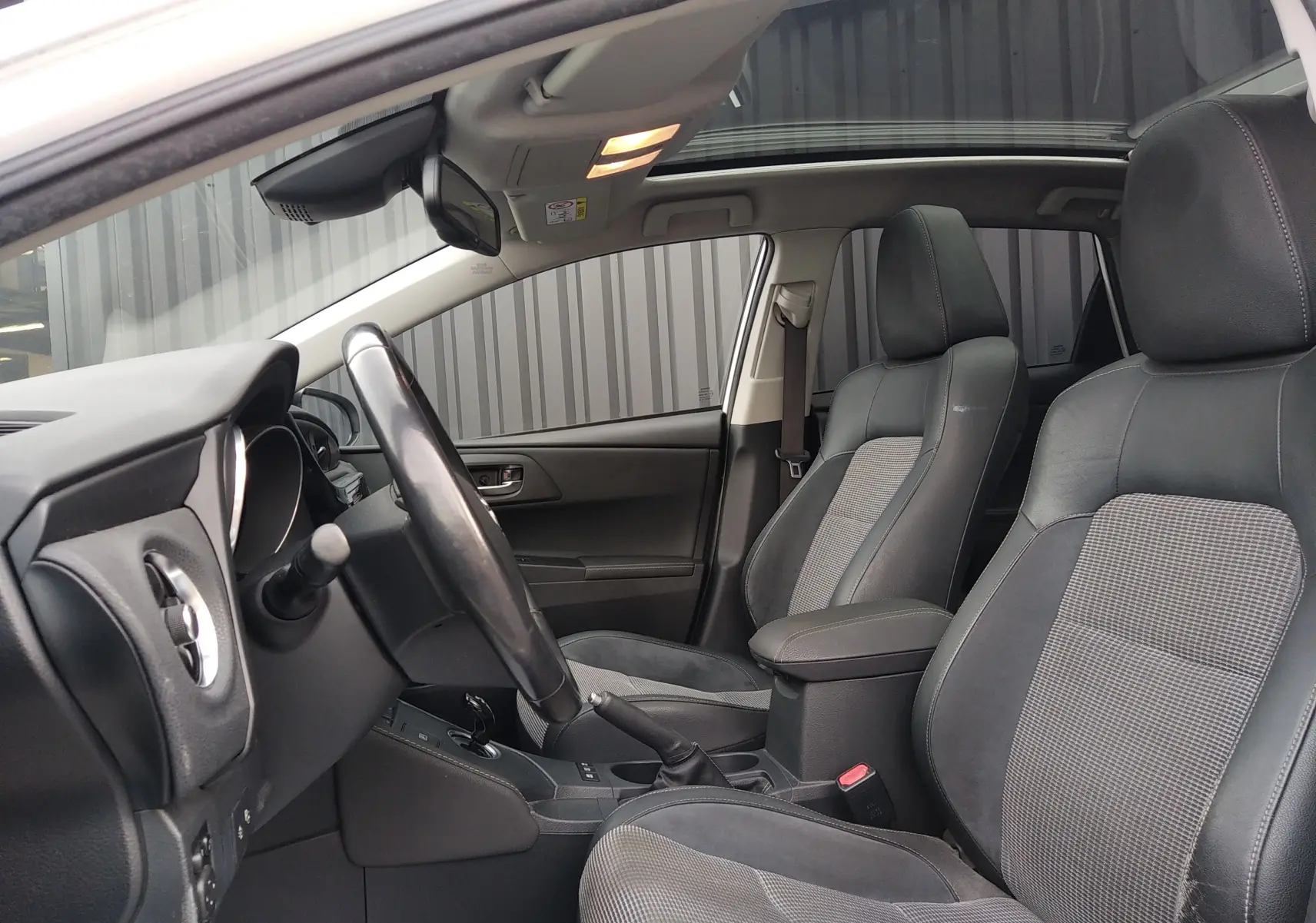 Vue intérieure côté conducteur de la Toyota Auris blanche, sièges en cuir noir avec inserts tissu gris et toit ouvrant panoramique.