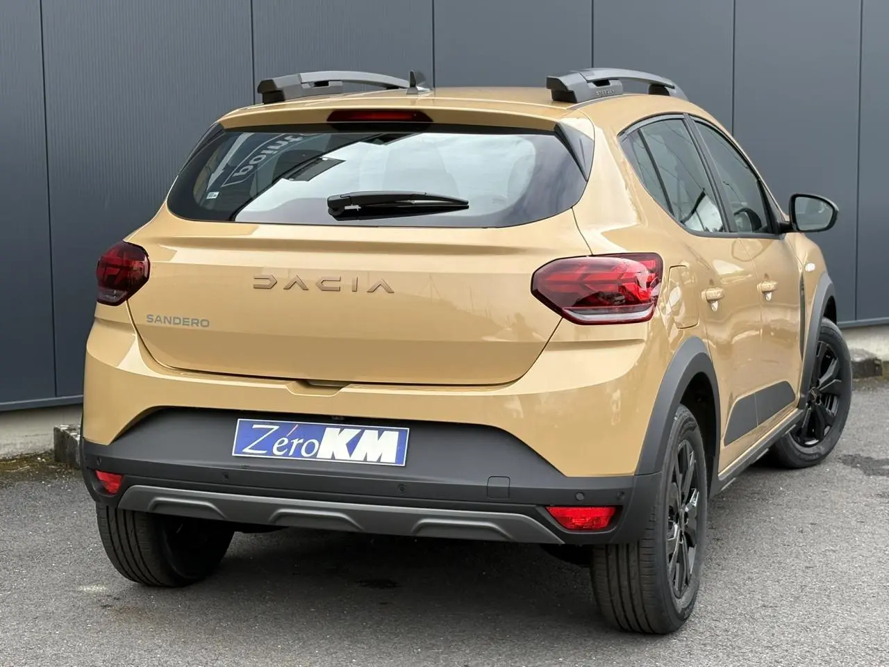 Vue 3/4 arrière droite d'une Dacia Sandero Stepway beige Safari avec barres de toit et jantes noires.