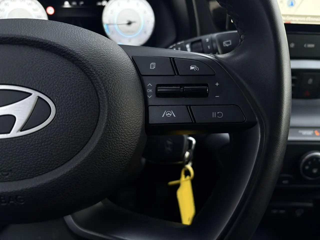 Gros plan sur le volant cuir noir de la Hyundai i20 2025 avec commandes au volant et compteur numérique flou en arrière-plan.