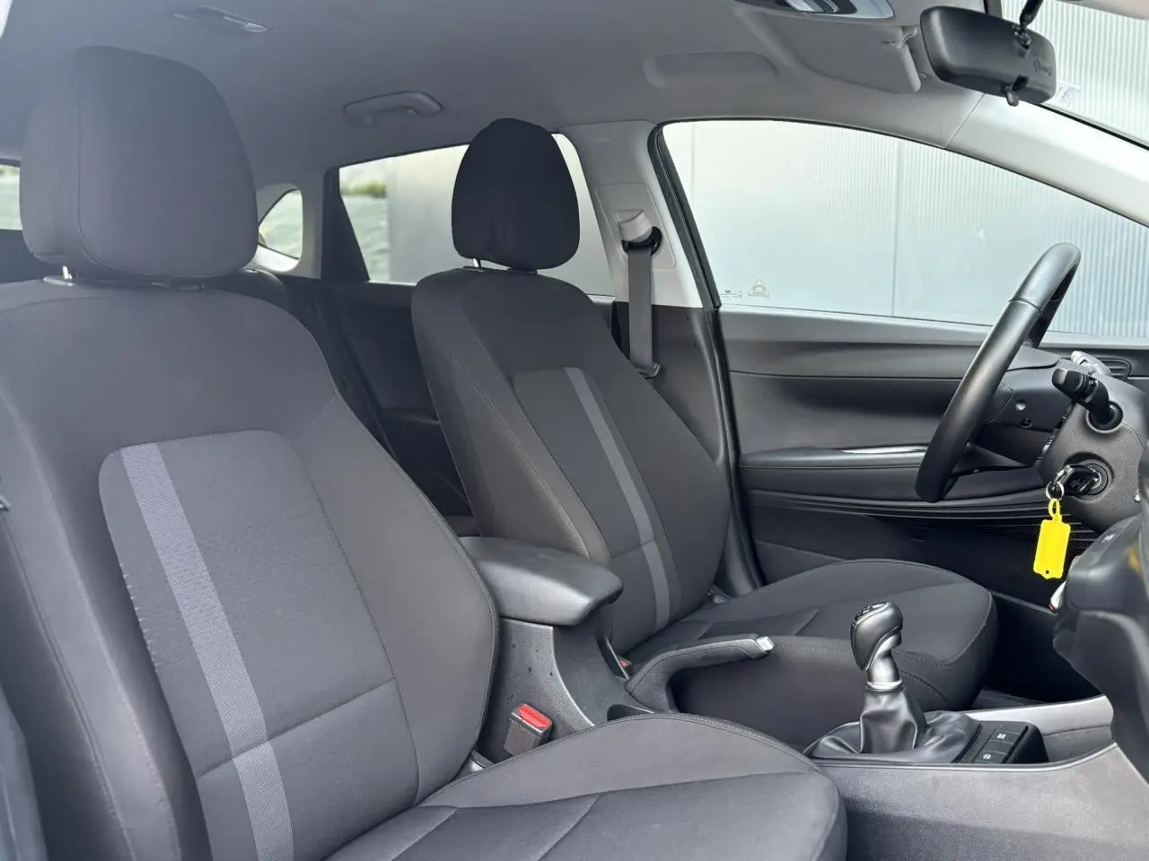 Vue intérieure côté droit de la Hyundai i20 2025, sièges tissu noirs avec accoudoir central et levier de vitesses manuel.